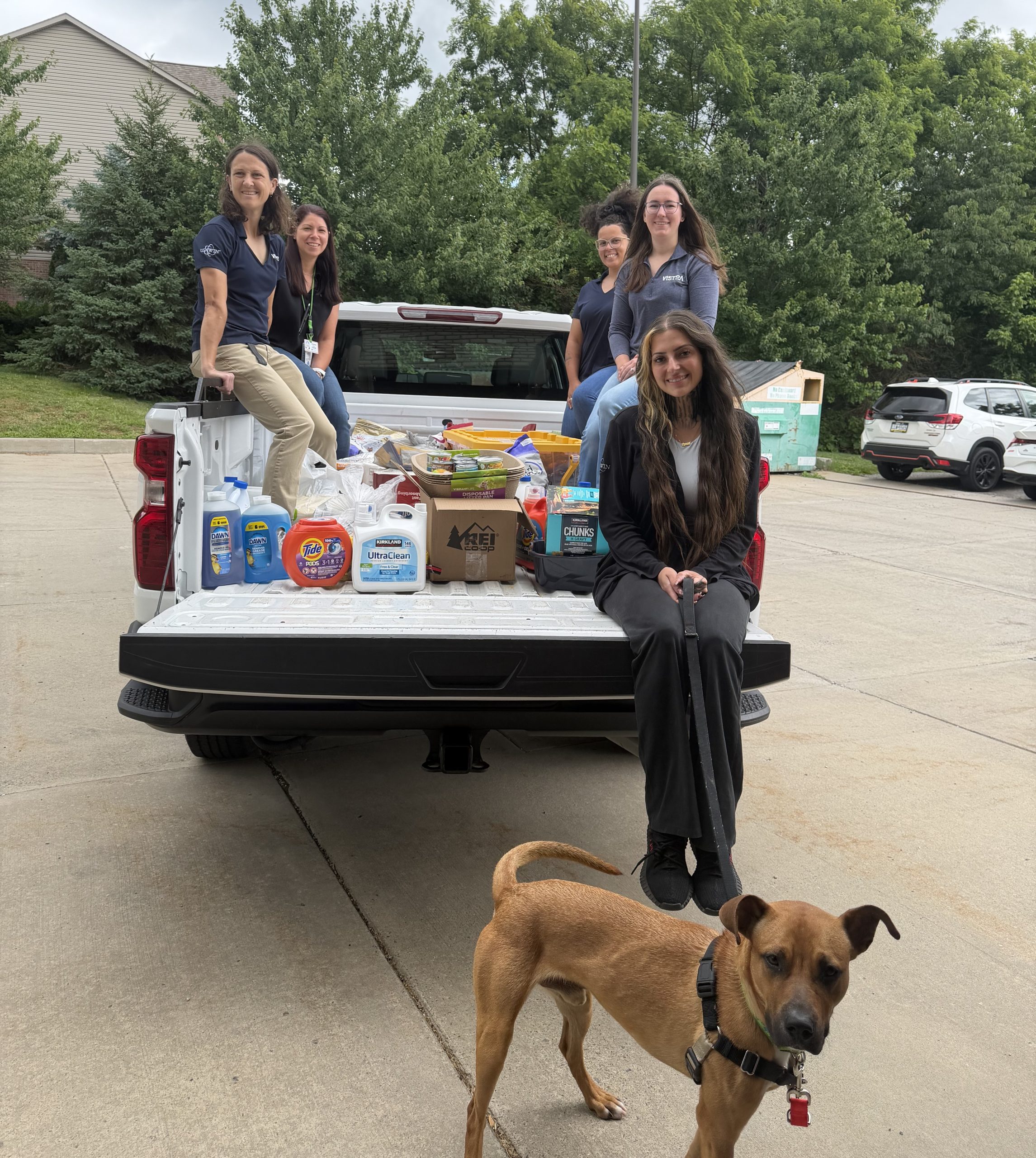 Five women and a dog in the foreground sitting on the back of an open pickup truck filled with shelter donations.